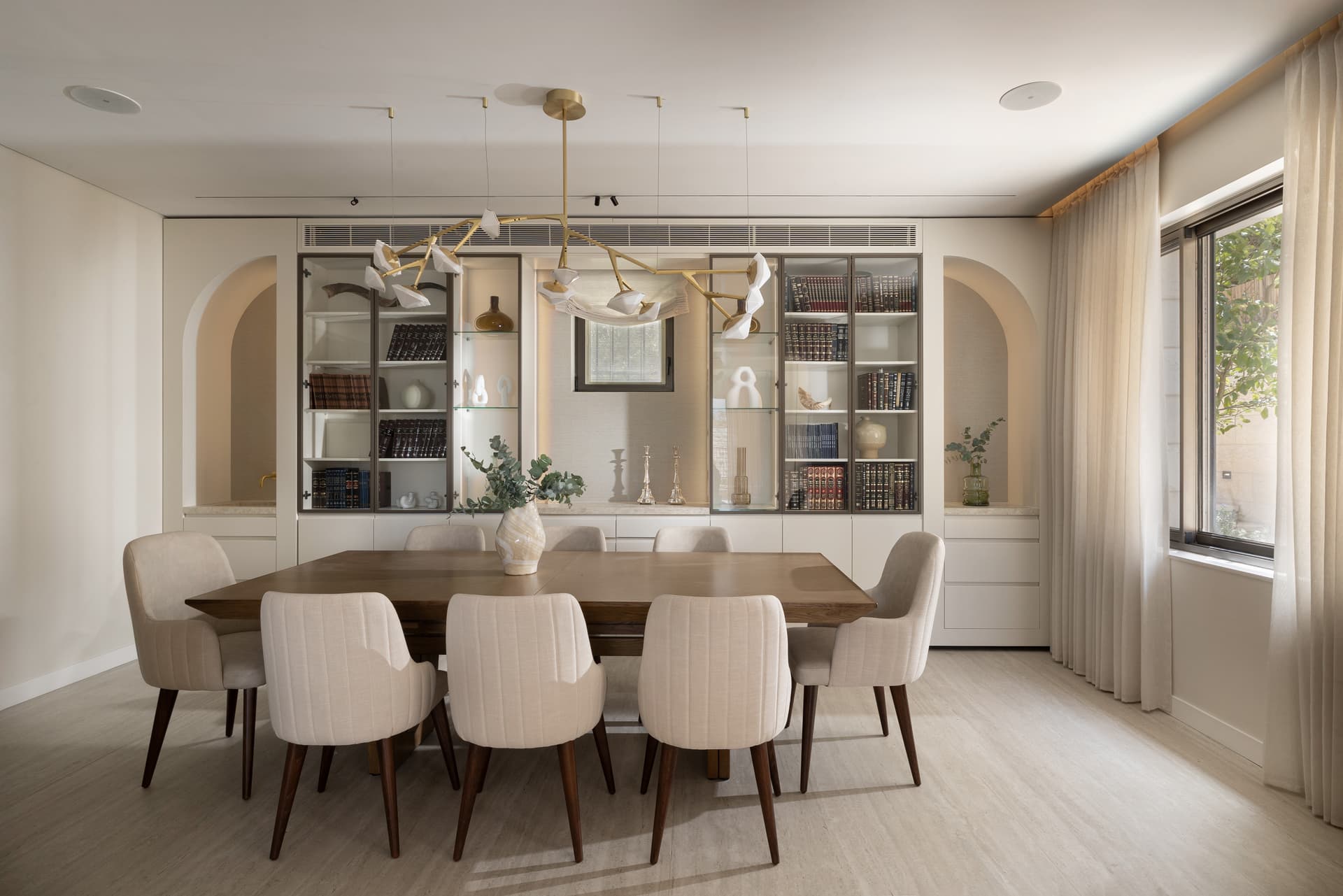 Formal dining room with walnut table, upholstered chairs, glass-front library wall, and brass branching chandelier