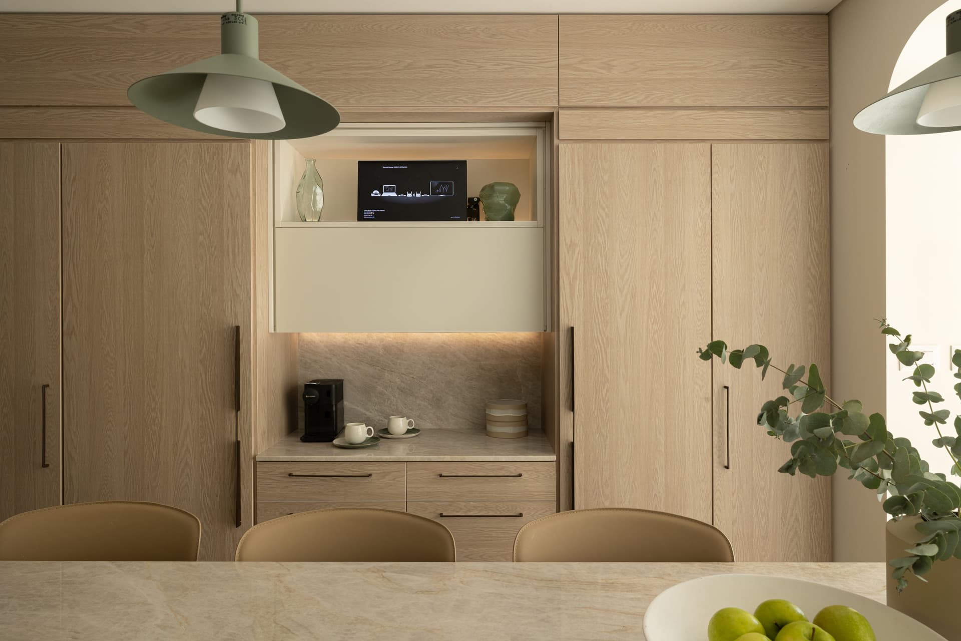 Kitchen seen through an oak doorframe, with sage island, pendant trio, and window seat beyond