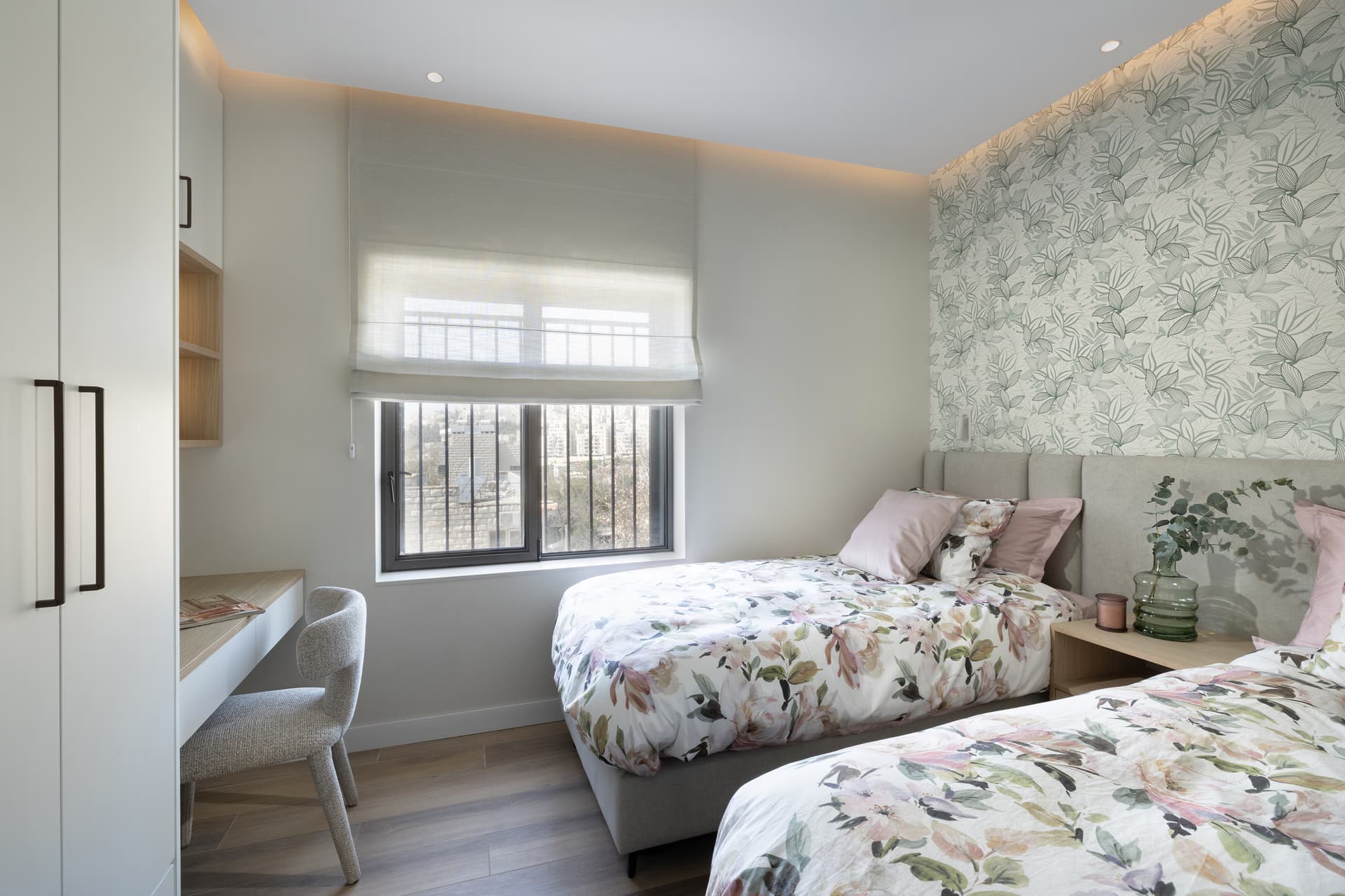 Children's bedroom with floral bedding, leaf-print accent wall, and a desk at the window with city views