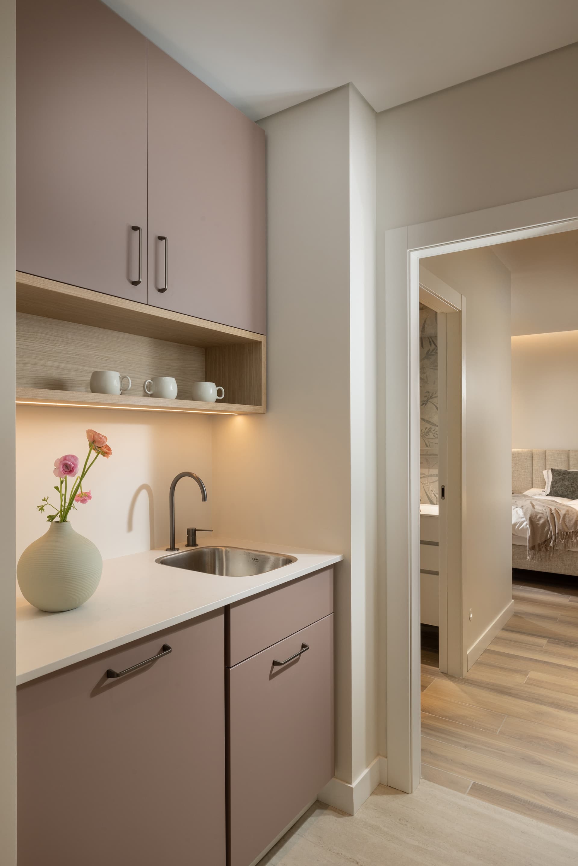 Dusty-pink kitchenette with brass tap, open oak shelf, and a view through to a bedroom